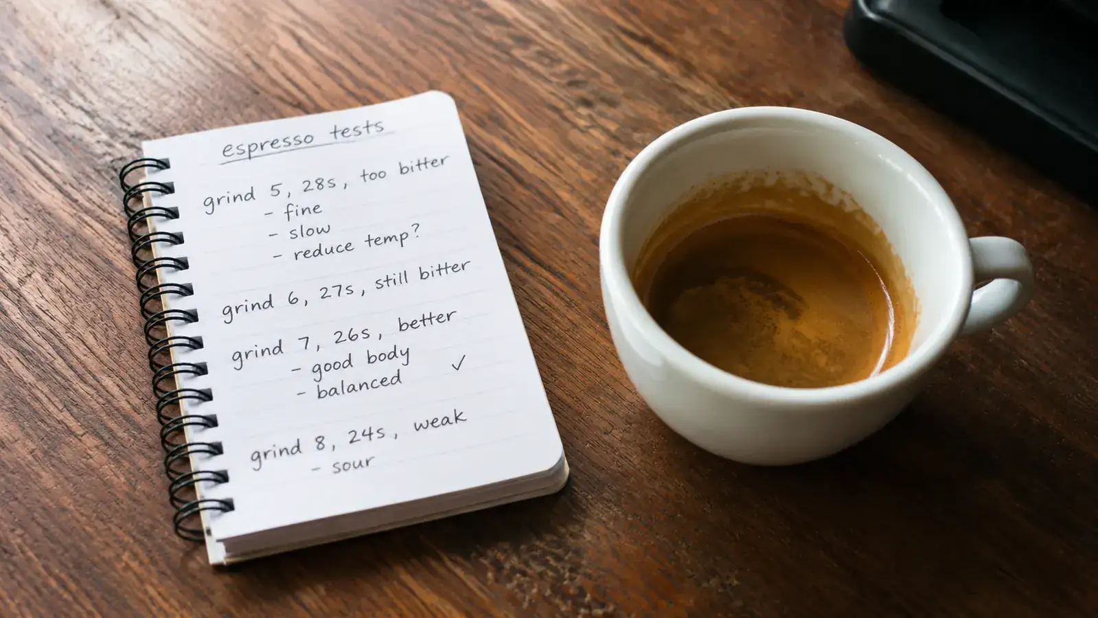 A notebook open on a kitchen table with handwritten grind settings and shot times, espresso cup beside it