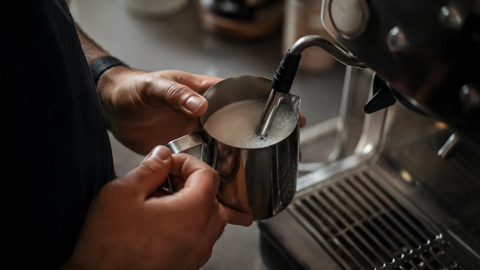 Person steaming milk in a small stainless pitcher at a home espresso machine on a kitchen counter