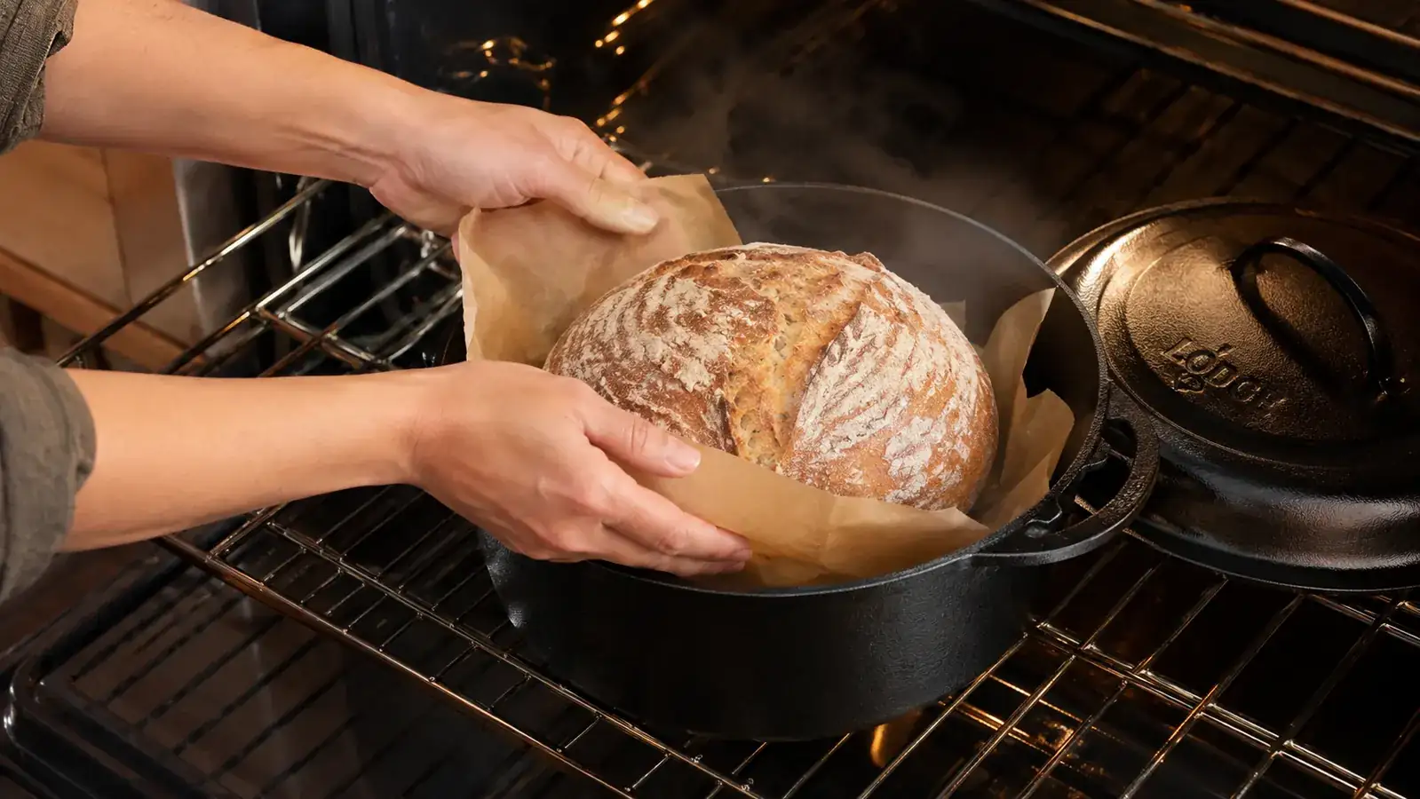 Hands lowering a shaped sourdough boule into a black cast iron dutch oven using a parchment sling